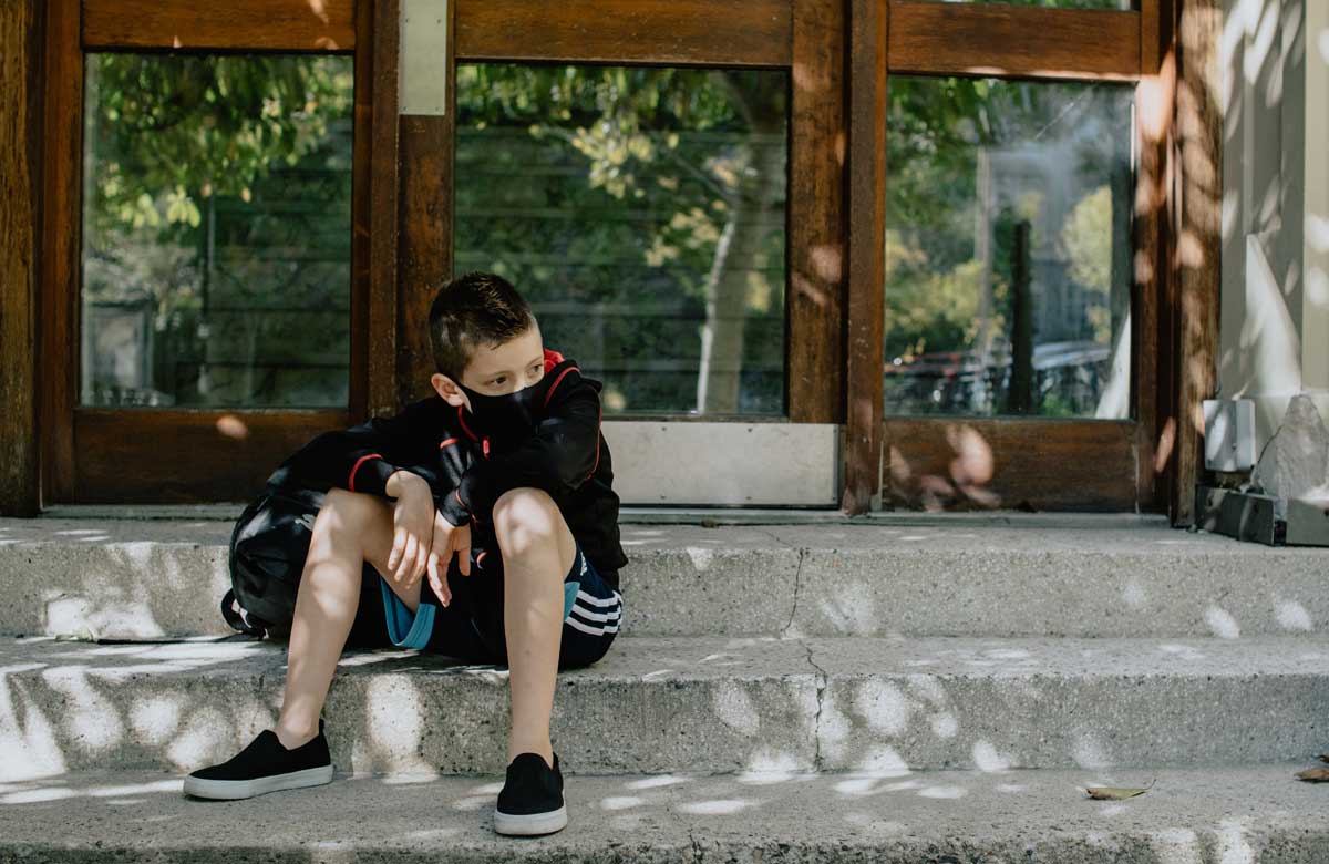 Child sits on steps with backpack, wearing a non-medical mask